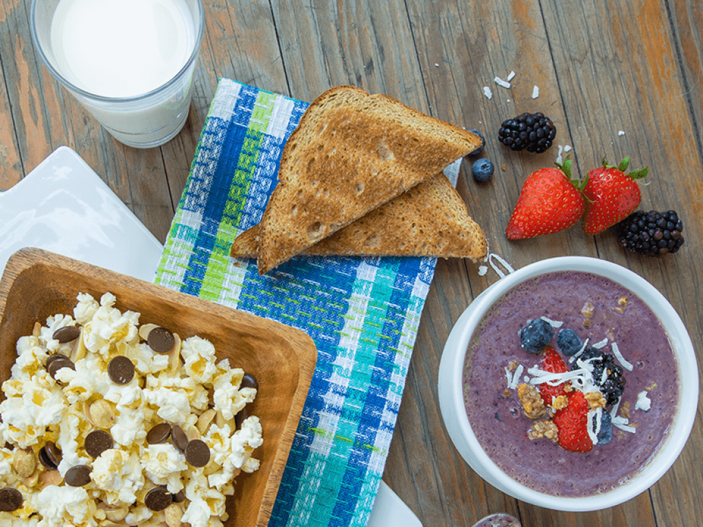 Aerial shot of a purple acai bowl with berries on top, next to a piece of toast and a bowl of popcorn mixed with chocolate chips.
