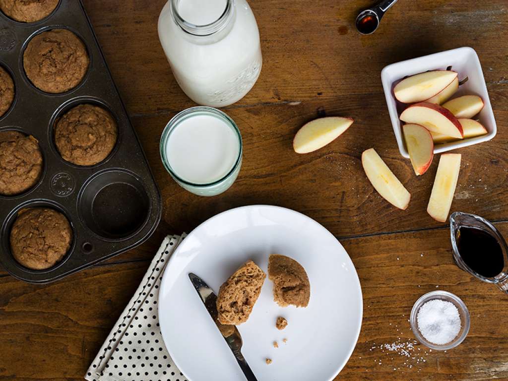 Birds eye shot of a cut muffin on a plate, neatly laid out next to a glass of milk, a jar of milk and sliced apples on a wooden table.