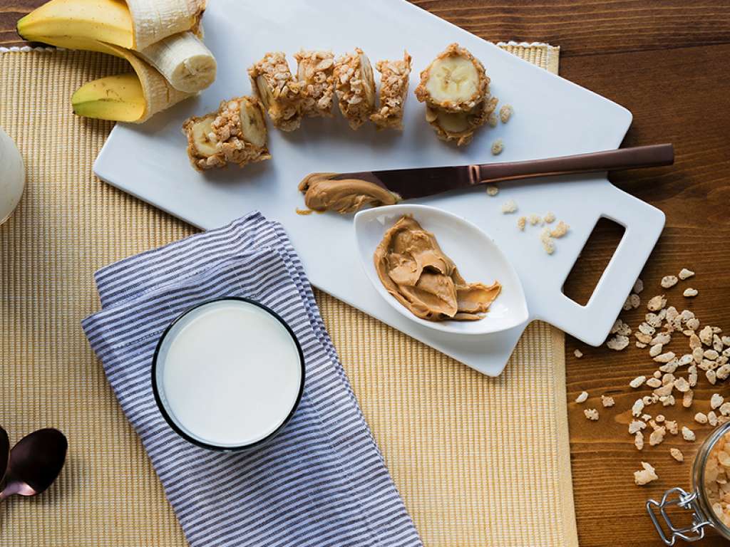 Aerial view of a banana, dipped in peanut butter and toped with coconut and nuts slices on a plate next to a glass of milk.