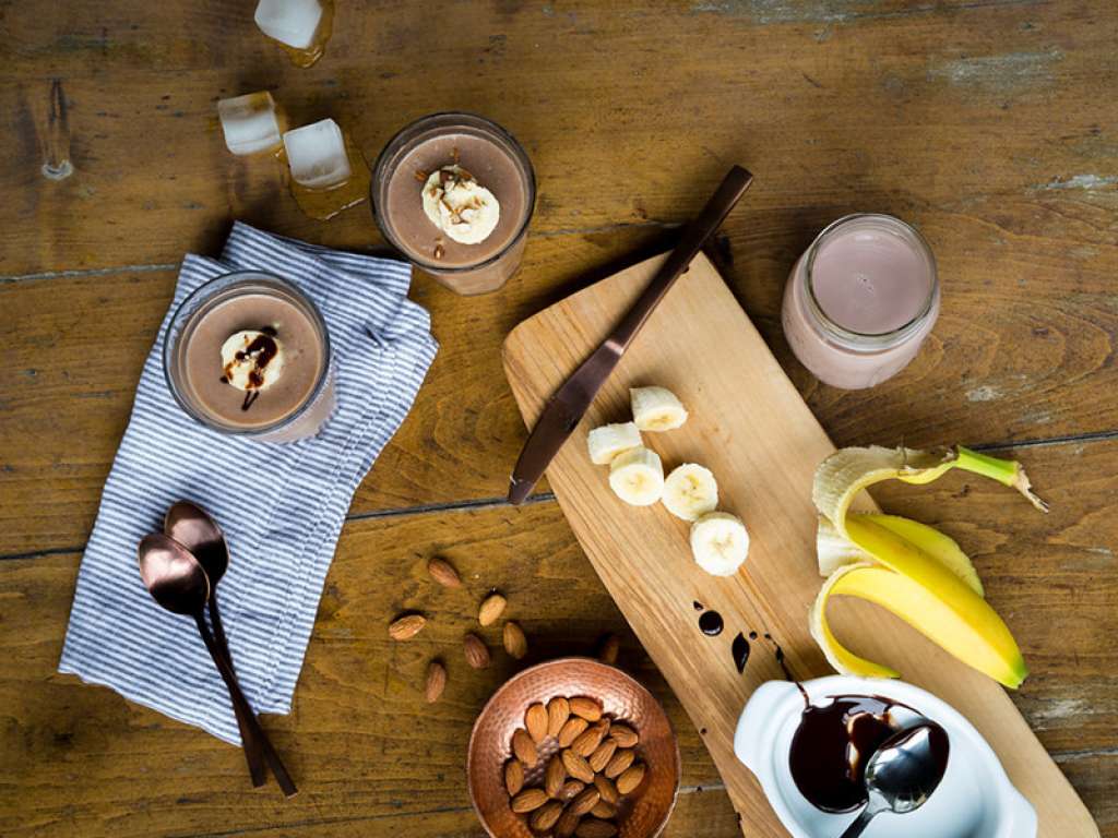 Aerial shot of two chocolate banana smoothies neatly surrounded by various ingredients, on a wooden tabletop.